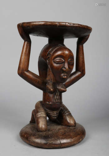 A West African carved hardwood figural stool. The circular seat is supported by a kneeling naked