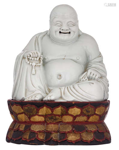 A Chinese porcelain figure of a smiling Budai, seated on a polychrome lacquered wooden lotus base, with his prayer beads resting on his raised knee, H 27,5 (without base ) - 40 (with base) - W 27,5 - D 24,5 cm