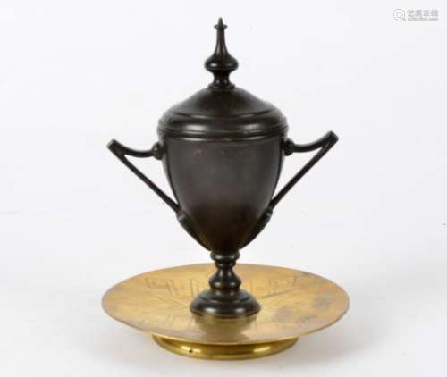 A bronze and brass inkwell, taking the form of a twin handled urn, raised on a geometric designed