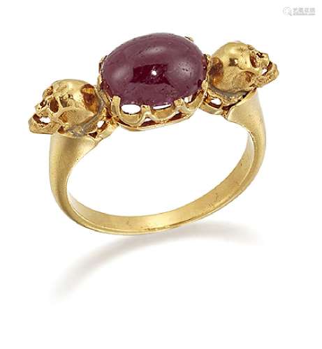 A ruby single stone ring, the oval cabochon ruby in claw -set mount between single skull motifs,