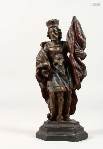 A 17TH/18TH CENTURY SMALL CARVED WOOD STANDING FIGURE, holding a flag, on a wooden base. 8ins high.