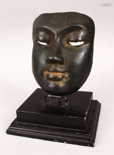 AN EASTERN BRONZE MASK with dark patination on a wooden base, 22cm high on stand, the mask 16cm high
