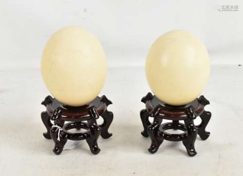 A pair of blown ostrich eggs mounted on Chinese hardwood stands, overall height 23cm, height of eggs