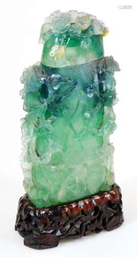 A green quartz vase and cover, heavily carved with flower heads etc., on a wooden plinth base,