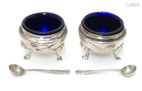 A pair of circular silver table salts, each raised on three hoof feet, with blue glass liners and