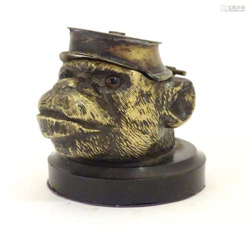 A Victorian novelty brass desk inkwell formed as the head of a monkey with glass eyes wearing a hat.