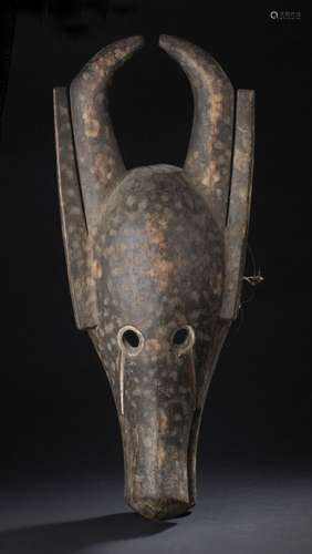 Masque zoomorphe à cornes, Bamana, MaliH. 61 cm L. 26 cmLe rythme de la courbe des cornes élancées se rejoignant en leurs sommets est accentué par la stricte linéarité et verticalité des oreilles qui y sont juxtaposées. La mâchoire plongeant vers le sol entrouverte s'élargit en son extrémité. Le front bombé, les yeux circulaires évidés révélés par un pourtour creusé. Patine tachetée alternance de brun clair et fonce.