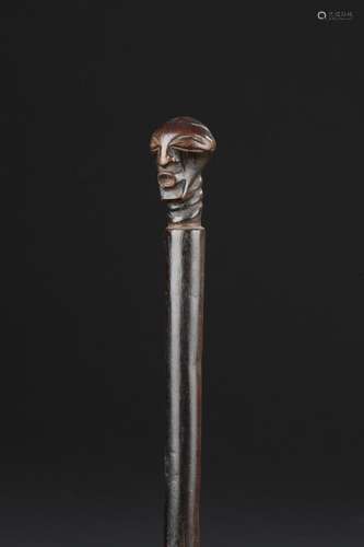 Sceptre de dignitaire songye, République Démocratique du CongoL.105 cmRare et bel insigne de pouvoir appartenant au roi, aux dignitaires, aux devins ou aux juges; véritable prolongement de la main, ce sceptre se compose d'une hampe cylindrique à la patine brune au nuances acajou, servant de support à une figurine, réincarnant très probablement un roi défunt. Cette effigie ancestrale est remarquable par l'expression du visage, et son raffinement épuré. Superbe traitement de la face polymorphe tendue vers l'avant, équilibrée. La rigueur de la stylisation géométrique mise en valeur par la finesse des traits aux arêtes saillantes, les orbites en reliefs ponctuées par les yeux étirés en amande. La calotte crânienne, en demi-sphère et léger rehaut. Très belle effigie qui s'impose, par sa remarquable qualité sculpturale, résumant à l'essentiel ses traits et conférant au visage une force puissante d'expressivité.