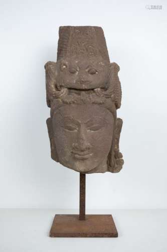 印度，11、12世纪。头戴高冠的神仙头像，装饰丰富，前面是张口吐珠的吉蒂穆卡。粉砂岩。高度：40厘米。缺少发型的顶部。一些震动和小缺憾。注：kirtimukha（梵文中的光荣面孔）是一个标志性的主题，经常用于建筑开口上方，但也作为装饰图案出现在佛龛或神灵的发型中，如这里。
