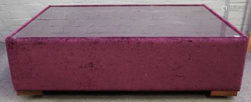 A large square plum coloured upholstered coffee table, with inset glass top on block supports,