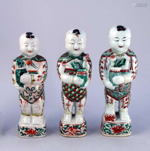 中国--康熙时期（1662-1722）。   三尊青花多色珐琅彩装饰的荷花瓷像，站在底座上，微笑着手持莲花瓶，衣服上饰有佛结、仙鹤、蜂巢等图案。   高度为25.7、27和27.5厘米。(小的缺口和磨损)