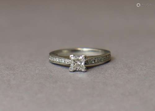 A DIAMOND RING, the princess cut stone set between a row of ...