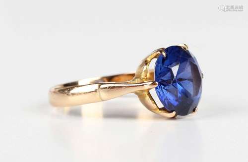 A gold ring, claw set with a circular cut synthetic sapphire...