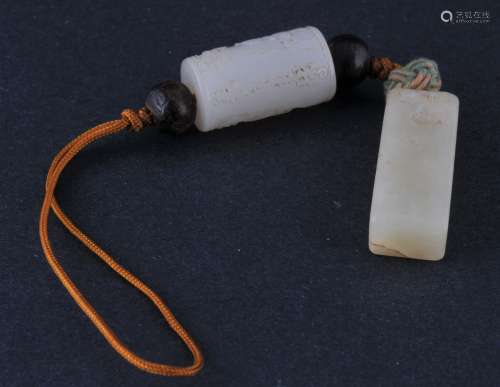 Two jade pendants. China. 19th century. A white jade cylinder carved with figures and a grey rectangle with inscriptions. Both threaded on the same cord.