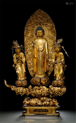 A GILT- AND BLACK-LACQUERED WOOD GROUP OF BUDDHA SHAKYAMUNI AND A PAIR OF BOSATSUS, Japan, Meiji period, Buddha Shakyamuni is standing in samabhanga on a separate carved lotus base placed on a cloud formation set on a rectangular base, both hands in vitarkamudra, wearing monk's garment falling down in pleats, his face displaying a serene expression, mandorla behind and flanked by a pair of bosatsus, one holding lotus flower, the other with hands in anjalimudra, both in bent posture, wearing dhoti, mantle and their faces displaying serene expressions - Property from a South German private collection, purchased from Kunsthandel Klefisch, Köln, Sale 13.01./02.12.1978, no. 559 - Partly small damages due to age and slightly rest.