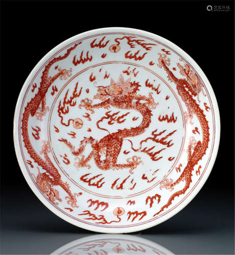 A RARE IRON-RED DECORATED 'DRAGON' DISH, China, underglaze-blue Kangxi six-character mark and period, painted in shaded tones of iron-red to the central medallion with a leaping five-clawed dragon chasing a flaming pearl, surrounded by pairs of striding dragons in mutual pursuit amid wispy flames in the cavetto and to the exterior, all enclosed by double-line borders