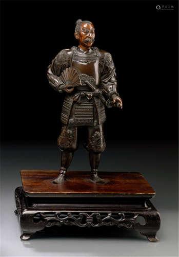 A BRONZE FIGURE OF A STANDING SAMURAI, Japan, Meiji period, cast in standing posture with fierce facial expression, wearing a robe with engraved floral patterns and crests, holding a fan in his right hand - German family property, acquired between 1960 and 1990 - Minor wear, sword attached to belt loose, mounted in wood stand