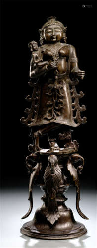 A BRONZE FIGURE OF A WOMAN CARRYING A CHILD, South-India, ca. 1900, standing with legs crossed on a high pedestal cast with four parrots, carrying a child in one arm, the other originally holding an attribute, wearing sari, bejewelled, her face displaying a serene expression with almond-shaped eyes and her hair combed in a long tress falling down her back - Property from a German private collection, collected between 1966 and 2004