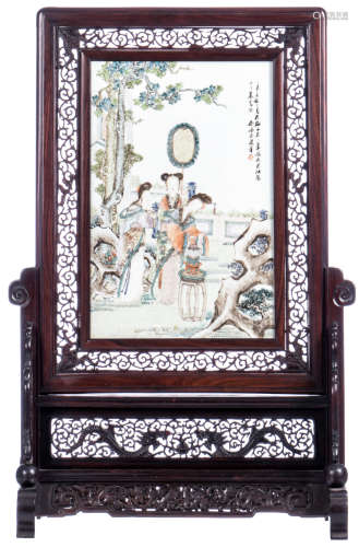 A Chinese richly carved wooden table screen, the porcelain plaque polychrome decorated with three ladies in a garden, signed, 19thC, H 70,5 - W 43,5 cm - 26 x 39 cm (plaque)