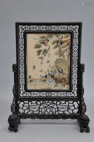 Table screen. China. 19th century. Embroidered silk panel with birds. Black wood frame carved and pierced with cash coins and archaic scrolling. Foo dog bases carved in the round. 23-1/2