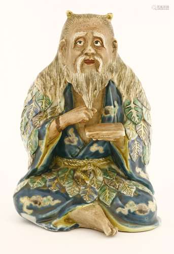 A Japanese Kutani figure, late 19th century, of a oni seated cross-legged with a book in his left ...