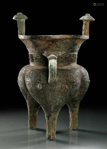A BRONZE TRIPOD VESSEL (JIA) FOR WINE OFFERINGS, China, Shang dynasty, 13th/ 12th ct. BC. With a tri-lobed body, tapering towards cylindrical legs, the wide flaring meck set with two studs with domed caps, the curved handle emerging from bull's head. The body is decorated with a taotie-motif on each bulb, the two eyes surrounded by deep spiralling lines. The neck shows a narrow band with further taotie-motifs with hooked eyebrows, centred on three raised flangs and extending into stylized bodies. The handle is plain except for a raised rib, the bull's head detailed with prominent eyes. The grooves of the design contain malachite encrustations. - Provenance: Bought in 2001 from L.H.W. Investment & Trading Limited, Hong Kong. - Described and illustrated in: Regina Krahl: Collection Julius Eberhardt. Early Chinese Art, vol. 2, Hong Kong 2004, p. 12f. For a related vessel see Zhongguo meishu quanji. Gongyi meishu bian: Qingtong qi, vol. 1, pl. 24 and Chen Peifen: Ancient Chinese Bronzes in the Shanghai Museum, London 1995, pl. 8.