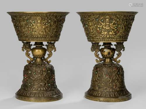 A PAIR OF BRASS BUTTER-LAMPS, Tibet, 19th ct., both standing on a domed-shaped base decorated with three multi-armed divinities seated on a lotus base in a medallion surrounded by scrolling tendrils, the containers ornamented to the outside with four multi-armed protective deities standing and seated on a lotus base placed in a medallion amidst scrolling tendrils issued by vases, both inlaid with beads, sealed - Property from a Rhineland-Palatinate private collection of Tibetan art - Minor wear