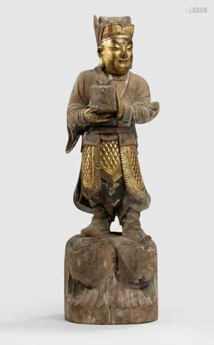 A WOOD FIGURE OF A YOUNG MAN, CHINA, Qing dynasty, standing on a base with both hands holding a box-shaped parcel, wearing various garments including a tunic and boots, his face displaying a smiling expression and his head covered with a hat, traces of gilt and red lacquer - Property from a Franconian private collection, bought 1995 - Minor damages due to age