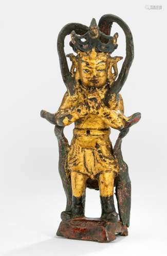 A GILT-LACQUERED AND PAINTED BRONZE FIGURE OF A STANDING GUARDIAN, China, Ming dynasty, standing on a pedestal with both hands clasped in front of the abdomen, wearing tunic, scarf and boots, his face displaying a severe expression and his head topped with a tiara - Property from an old North German private collection - Stand partly with losses, wear