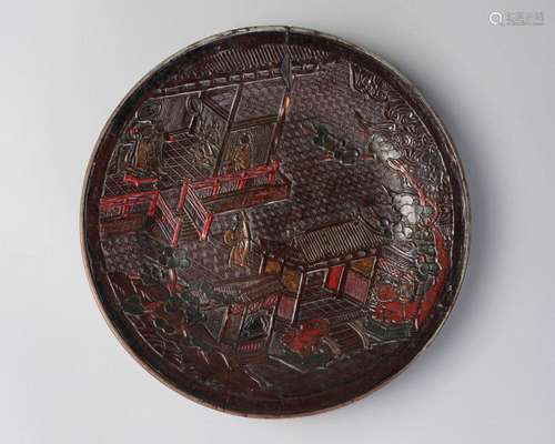 A Chinese lacquer plate, 18th century, carved and coloured with figures in a pagoda and courtyard, guarded by a pair of Buddhist lions, and surrounded by pine trees, 29cm diameter, with paper label inscribed 'W. R-S. Col'