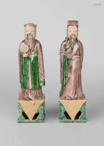 A pair of Chinese biscuit porcelain officials, Kangxi period, one modelled holding a leaf, the other a fly whisk, their robes glazed in aubergine and green, on pierced rectangular bases, 25cm high