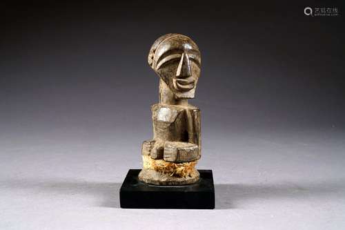 Fétiche Songye. La tête tournée vers la gauche, les deux mains posées sur l'abdomen au nombril saillant. Bois sculpté et patiné. République démocratique du Congo.<br/>Hauteur: 17 cm. Sur socle.