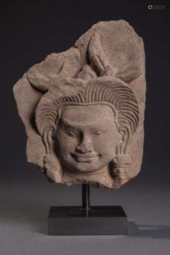 Haut relief de frise de sanctuaire ciselée d'une tête de Devata, la coiffure à longues mèches rayonnantes surmontées de plumes hérissées, paré de lourds pendants d'oreilles, le sourire de béatitude typique.