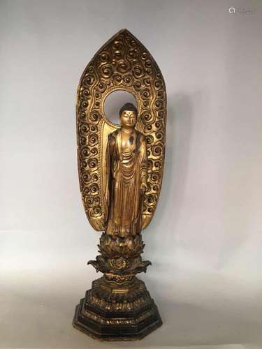 Buddha Amida Nyoraï, forme du Buddha Amitabha debout sur un haut tertre élaboré terminé par un lotus à plusieurs pétales épanouis dans une posture hiératique vêtu de sa robe monastique plissée recouvrant ses deux épaules et retombant sur ses pieds, marquant de sa main droite le geste de l'argumentation Virtaka Mudra, coiffé de fines bouclettes, orné d'un joyau à la base de la protubérance crânienne ushnisha, le visage empreint de beauté et sérénité.