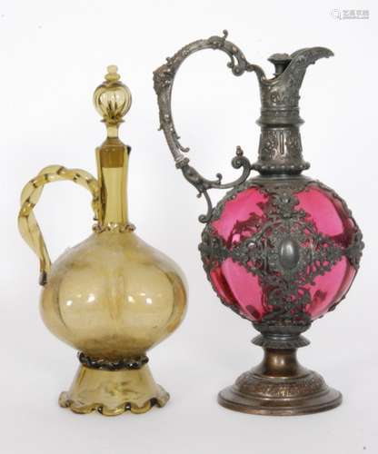 An early 20th Century Continental glass claret jug, conical foot rising to the lobed body below a shaft neck with applied rope twist handle and hollow blown stopper all to an olive green ground, unmarked, height 33cm, together with  a another example with a cranberry glass body encased by white metals mounts and foot with scrolling decoration between cartouche panels, unmarked, height 34cm. S/D (2)