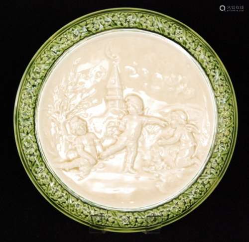 An early 20th Century Arnold and Ludwig Richard Schutz majolica charger decorated in relief with a scene of three winged elves playing in a garden under a moonlit sky, all in cream with a green moulded border egde, impressed Schutz Cilli mark, diameter 38.5cm