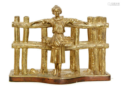 A late 19th Century brass letter rack modelled as a standing country girl leaning with her back to a fence, all to a wooden base, length 33cm.