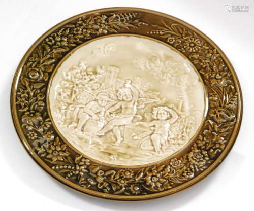 An early 20th Century Arnold and Ludwig Richard Schutz majolica charger decorated in relief with a scene of three winged cherubs playing in a castle garden under a moonlit sky, all in cream with a brown moulded floral border edge, impressed Schutz Cilli mark, diameter 45.5cm