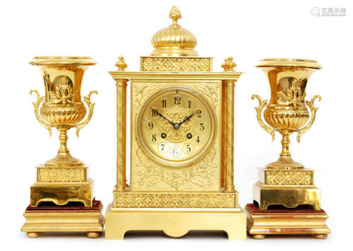 A late 19th Century eight day strike gilt mantle clock garniture, the dial with Arabic numerals flanked by four column supports below a dome pediment and flame finial, height 35cm, with associated side twin campana urns raised on a wooden base.