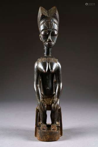 Figurine Baoulé. Représentant une jeune femme assise sur une chaise, les mains posées sur les genoux. Le dos, les épaules et le cou, parcourus de scarifications. Elle est vêtue d'un pagne noué à la taille. Beau visage allongé à la coiffe bipartite sophistiquée et aux yeux clos. Bois sculpté à patine sombre. Côte d'Ivoire. Hauteur: 46 cm. Provenance: ancienne collection de Madame J. de L. à Bruxelles. Acquise dans les années 70 à Monsieur Emile Deletaille. Vendu pour cause de succession au profit de diverses associations.