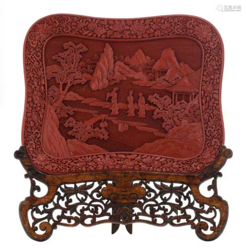 A Chinese carved red cinnabar plate, depicting courtladies in a garden, with a matching wooden stand, H 32,9 - W 39,5 - D 4,6 cm