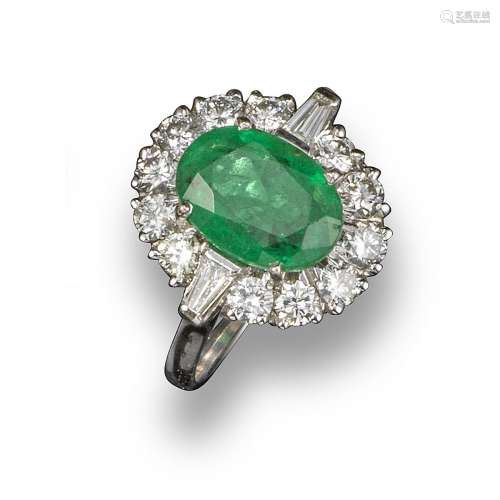 An emerald and diamond cluster ring, the oval-shaped emerald set within a surround of twelve