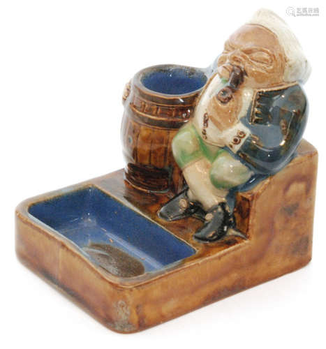 An early 20th Century Royal Doulton Lambeth vesta and ashtray formed as a seated man smoking a pipe with his arm around a barrel