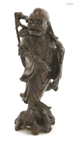 A Chinese wooden figure, c.1900, of a monk in a long robe, carrying a fly whisk on his shoulder and ...