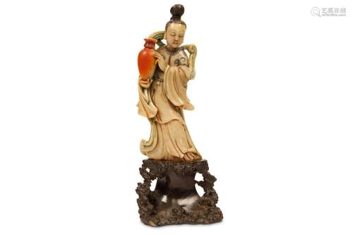 A CHINESE SOAPSTONE CARVING OF A LADY. Kangxi. Standing in long flowing robes with trailing ribbons swirling over the shoulders, a red vase supported in the right hand, the hair raised in a high bun, standing on an attached rockwork base, traces of