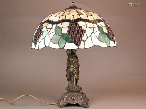 Table lamp in Tiffany style - bronze cast metal, curved foot with relief decoration, shaft<br />as a plastic caryatid, shade made of colored glass with lead bands, Art Nouveau motifs with stylized grape motif, 1-flame electrified, functions, H.ca.54cm.