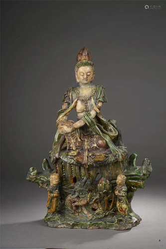 Le Boddhistava Kwan Yin   assis en délassement sur un haut tertre moulé   à la base de deux attendants un dragon et un lotus,   richement vêtu et paré de   joyaux. Céramique vernissé   sancaï, beige, brune et verte. Chine. Dynastie Ming. 1368 à 1644. Ht 46 x 34cm. Restauration. Test de thermoluminescence   confirmant la datation.