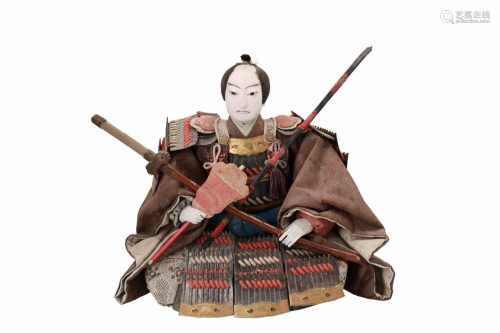 A doll of a sitting Japanese samurai warrior, surrounded with arrow feathers on a rack and a bow.